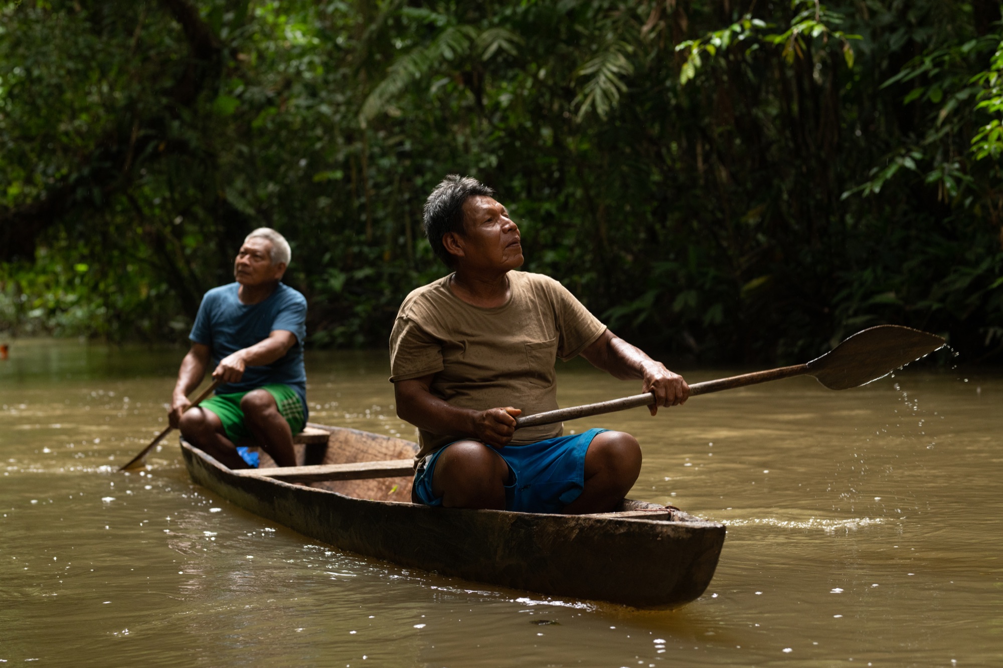 Elders paddle a canoe along a forest river.